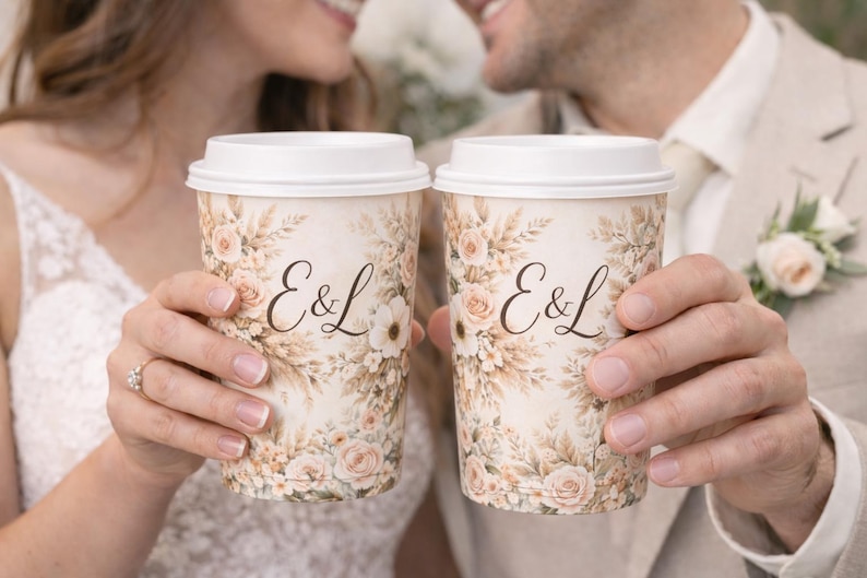 May include: Two coffee cups with white lids are held up, each displaying the initials "E&L" in elegant script. The cups are adorned with a floral design in soft, muted tones, suggesting a wedding or celebratory theme.