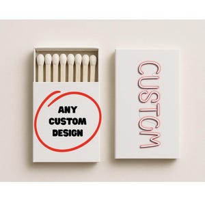 May include: Two white matchboxes. One box is open, revealing matches with white tips. The other box has the word "CUSTOM" in pink lettering. The open box has the text "ANY CUSTOM DESIGN" inside a red circle.