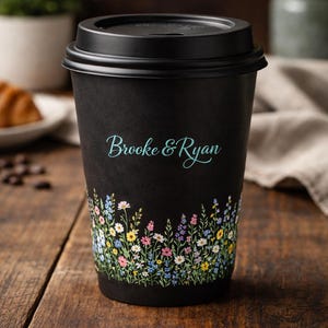 May include: Black disposable coffee cup with a floral design at the bottom and the names "Brooke & Ryan" in teal script. The cup is on a wooden surface.