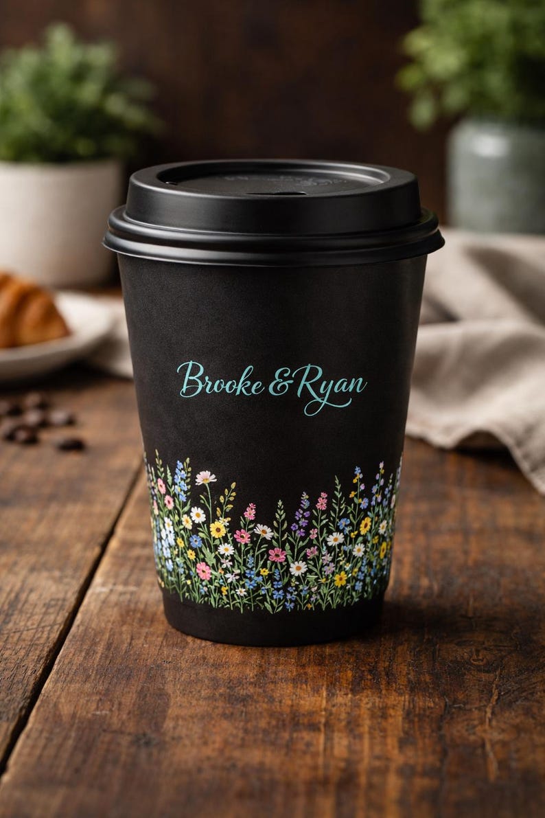 May include: Black disposable coffee cup with a floral design at the bottom and the names "Brooke & Ryan" in teal script. The cup is on a wooden surface.