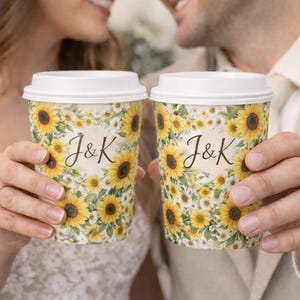 May include: Two coffee cups with white lids, decorated with sunflowers and daisies. The cups have the initials "J&K" in elegant script. The cups are held by two people, suggesting a wedding or special occasion.