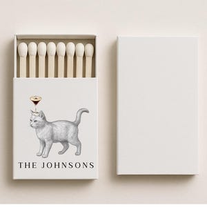 May include: A white matchbox with a gray cat illustration wearing a cocktail glass on its head. The text "THE JOHNSONS" is below the cat. The box is open, revealing matches. A separate, blank white box is to the right.