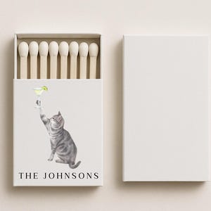 May include: A white matchbox with a watercolor illustration of a gray tabby cat reaching for a margarita. The text "THE JOHNSONS" is printed below the cat. The matchbox is open, revealing wooden matchsticks, and is next to a closed, blank white box.
