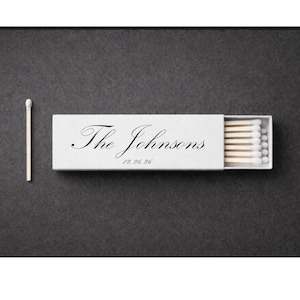 Pode incluir: Uma caixa de fósforos branca com o texto "The Johnsons" em escrita elegante e uma data. A caixa está parcialmente aberta, revelando fósforos com pontas brancas. Um fósforo solitário está ao lado.
