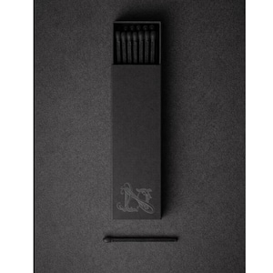May include: A black matchbox with a decorative emblem and a single match. The box is open, revealing several black matches inside. The image is monochromatic, with a focus on the texture and design of the matches and box.