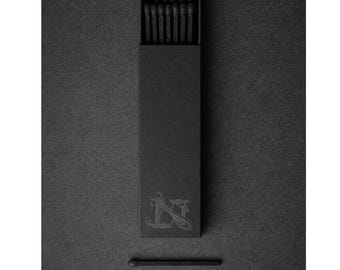 May include: A black matchbox with a decorative emblem and a single match. The box is open, revealing several black matches inside. The image is monochromatic, with a focus on the texture and design of the matches and box.