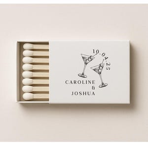 May include: A white matchbox with a partially open drawer revealing wooden matches. The box is decorated with two martini glasses and the names "Caroline & Joshua" with a date above. The matches are arranged neatly.