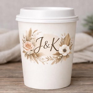 May include: White paper coffee cup with a white lid. The cup is decorated with a floral design in neutral tones, including roses, pampas grass, and the initials "J&K". The cup is sitting on a wooden surface.