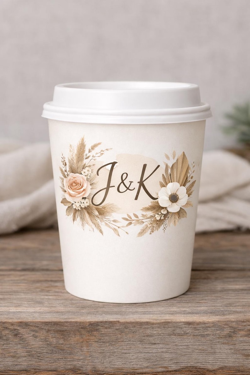 May include: White paper coffee cup with a white lid. The cup is decorated with a floral design in neutral tones, including roses, pampas grass, and the initials "J&K". The cup is sitting on a wooden surface.