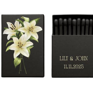 Puede incluir: Caja de cerillas negra con un diseño floral de lirios blancos y hojas verdes. La caja de cerillas abierta revela cerillas negras. El texto "LILY & JOHN" y la fecha "11.11.2025" están impresos en dorado.