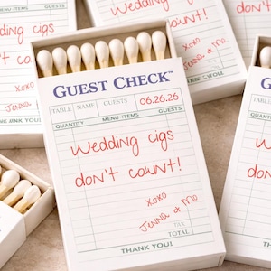 May include: White matchboxes with the text "GUEST CHECK" and the date "06.26.26". Handwritten text in red says "Wedding cigs don't count!" and "Xoxo Jenna & Mo". Each box contains white-tipped matches.