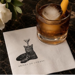 May include: A white paper napkin featuring a black and white illustration of a cat with a martini glass balanced on its head, accompanied by the text DRINKS ON COWBOY, positioned next to a glass of amber liquid with a large ice cube and orange peel.