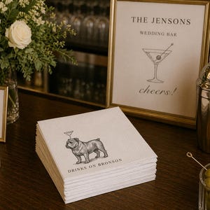 May include: A stack of white paper napkins with a bulldog illustration and the text "DRINKS ON BRONSON". The napkins are on a dark wooden surface, near a framed sign that reads "THE JENSONS WEDDING BAR".