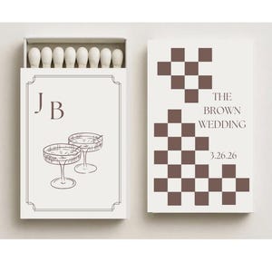 Op de afbeelding: Een crème-kleurige luciferdoos met een bruin geblokt ontwerp en de tekst "THE BROWN WEDDING 3.26.26". De voorkant van de doos toont de initialen "JB" boven een schets van twee champagneglazen.