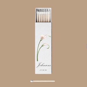 May include: A white matchbox with a floral design featuring calla lilies. The box is labeled "Johnsons" with a date below. A single match is placed below the box on a tan background.