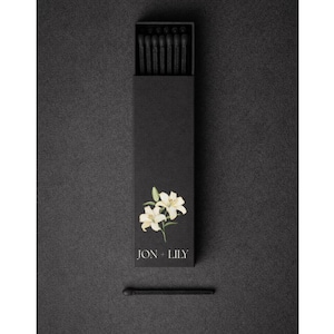 May include: A black matchbox featuring a floral design and the text "JON + LILY". The box is open, showing black matches. A single match lies below the box on a dark surface.