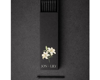May include: A black matchbox with a floral design and the text "JON + LILY". The box is open, revealing black matches. One match is placed separately below the box on a dark surface.