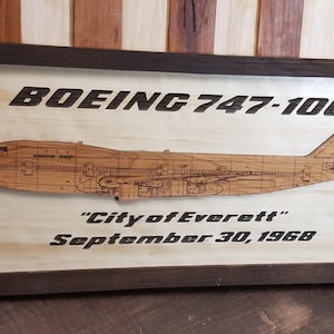 May include: A wooden plaque with a dark brown frame features a laser-cut design of a Boeing 747-100 airplane. The plaque reads "Boeing 747-100 "City of Everett" September 30, 1968".
