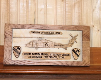 May include: Wooden framed plaque featuring a laser-cut Sikorsky UH-60A Black Hawk helicopter. The plaque includes text identifying the aircraft and the Combat Aviation Brigade, 1st Cavalry Division, 7th Squadron, Fort Cavazos, Texas. Two emblems are also included.
