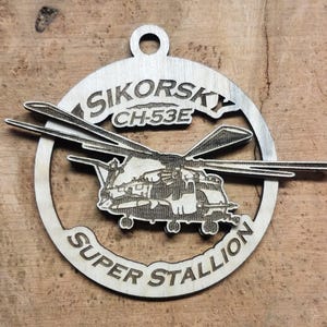 May include: A wooden ornament featuring a black and white silhouette of a Sikorsky CH-53E Super Stallion helicopter. The ornament is round and has a hole at the top for hanging.