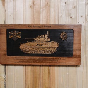 May include: A rectangular wooden plaque featuring a laser-engraved Bradley fighting vehicle in the center, flanked by a heart-shaped emblem and a Native American headdress icon. The text SSG Sterling T. Bienek is engraved at the top of the frame.