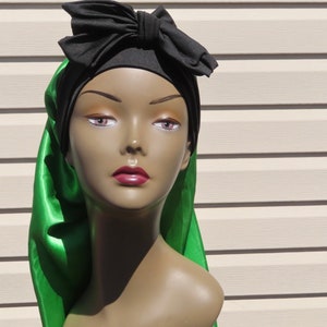 Pode incluir: Um lenço de cabeça preto com um grande laço decorativo na frente, combinado com uma cauda longa e fluida de tecido verde acetinado que desce pela lateral. O lenço é exibido numa cabeça de manequim com traços faciais definidos.