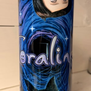 May include: A blue tumbler featuring the word "Coraline" in a stylized font, along with illustrations of a character and a black cat with blue eyes. The design includes bats, stars, and a spiderweb.