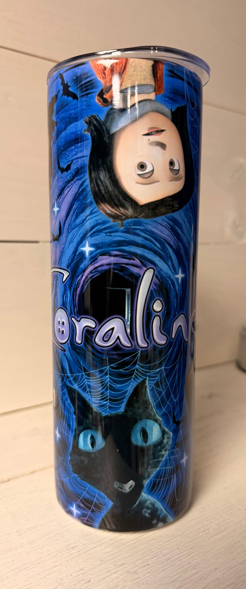May include: A blue tumbler featuring the word "Coraline" in a stylized font, along with illustrations of a character and a black cat with blue eyes. The design includes bats, stars, and a spiderweb.