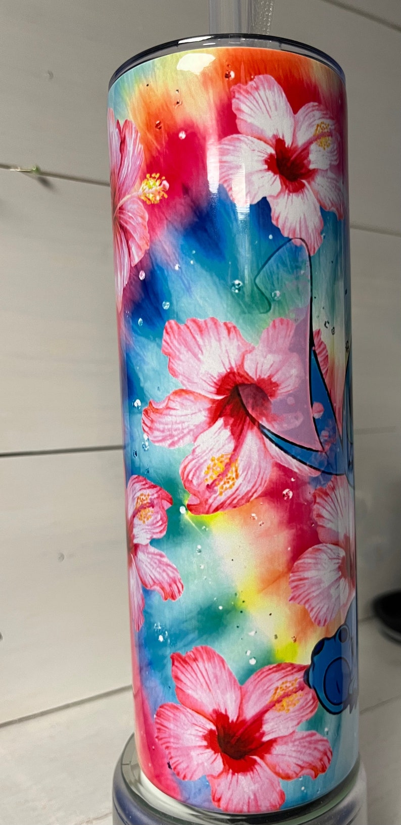 Puede incluir: Un vaso de acero inoxidable con un dise&ntilde;o de tie-dye colorido con flores de hibisco rosas y un personaje de dibujos animados azul.