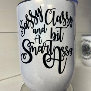May include: White tumbler with a black glitter finish and black lettering that reads "Sassy Classy and a bit Smart Assy".