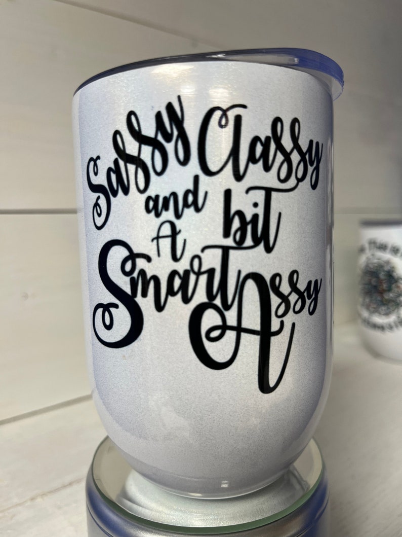 May include: White tumbler with a black glitter finish and black lettering that reads "Sassy Classy and a bit Smart Assy".
