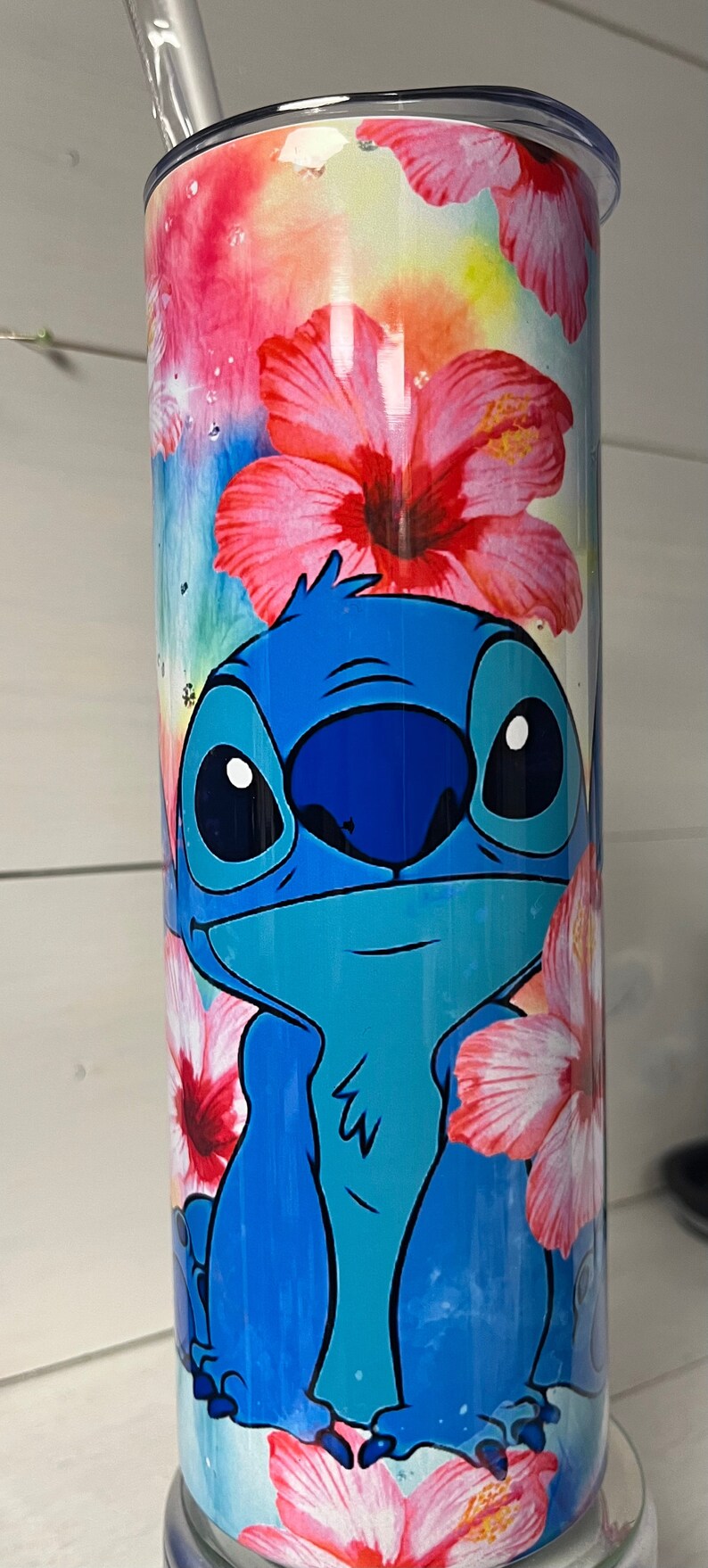 Puede incluir: Un vaso de acero inoxidable con un dise&ntilde;o floral colorido en rosa, amarillo y naranja. El personaje de Stitch, azul y turquesa, de Lilo & Stitch est&aacute; representado en el vaso.