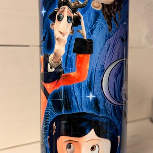 May include: A cylindrical tumbler featuring characters from the animated film "Coraline." The design includes illustrations of Coraline, the Other Father, and other characters against a blue background with spiderwebs and stars.