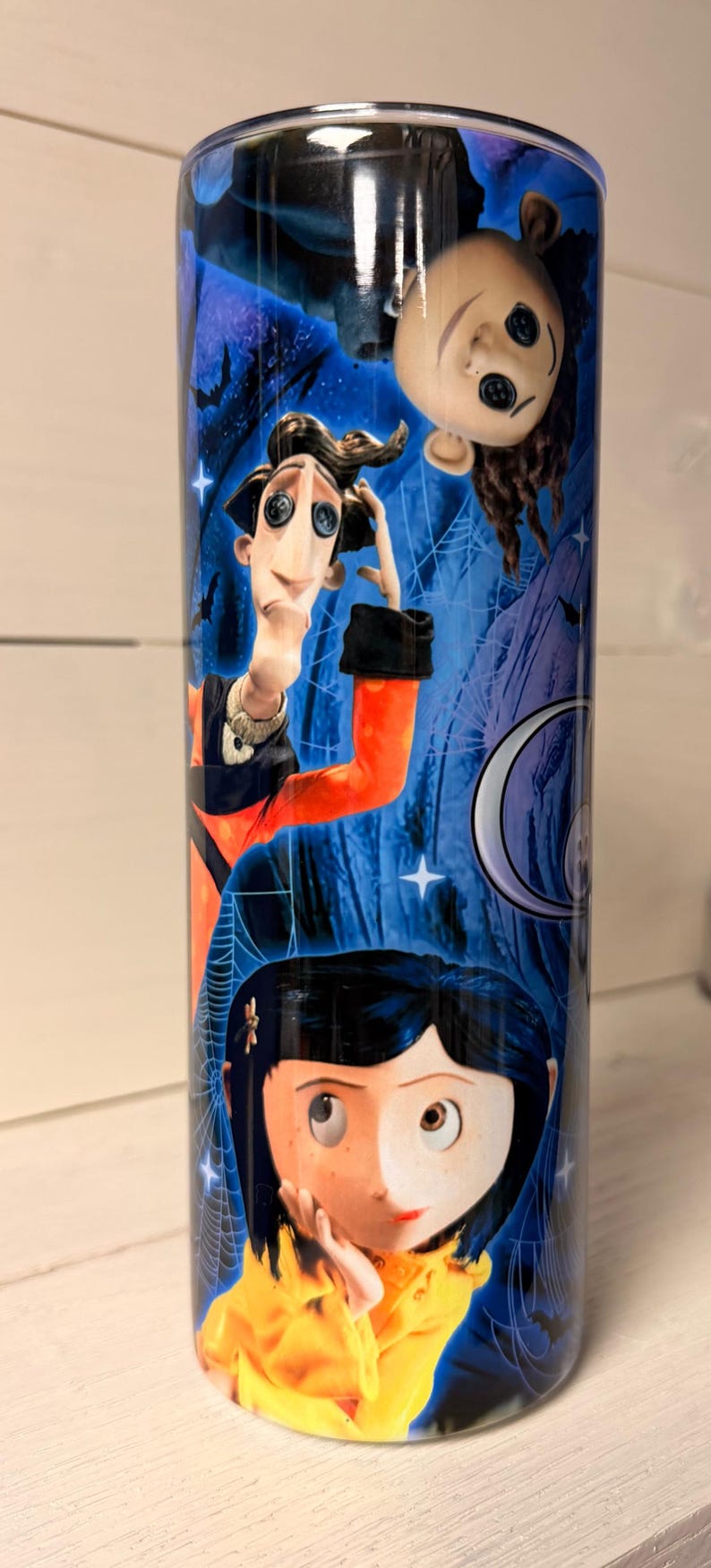 May include: A cylindrical tumbler featuring characters from the animated film "Coraline." The design includes illustrations of Coraline, the Other Father, and other characters against a blue background with spiderwebs and stars.
