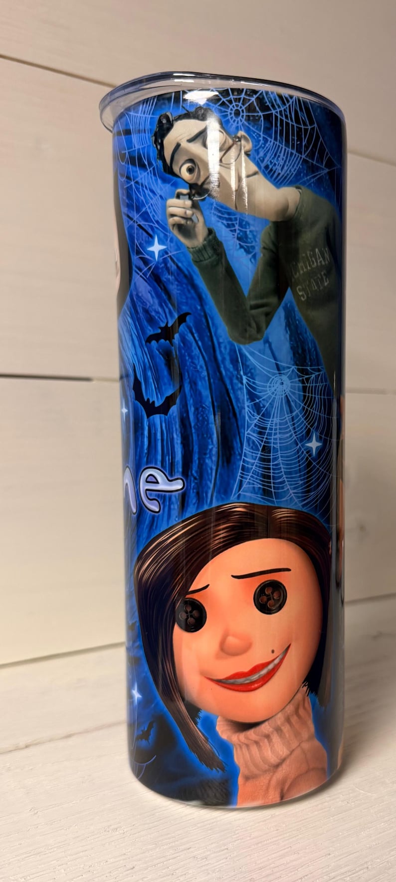May include: A stainless steel tumbler featuring characters from the animated film "Coraline." The design includes images of Coraline, the Other Father, bats, and spiderwebs against a blue starry background. The tumbler has a black lid.