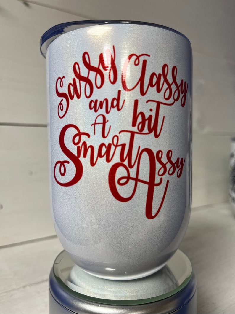 May include: White tumbler with a red glitter finish and red vinyl lettering that reads "Sassy Classy and a bit Smart Assy".