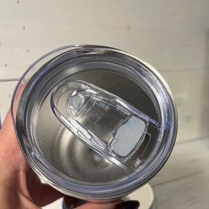 May include: Close-up of a clear plastic lid for a stainless steel tumbler. The lid has a silicone ring around the edge and a flip-top lid with a small handle.