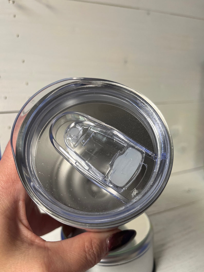 May include: Close-up of a clear plastic lid for a stainless steel tumbler. The lid has a silicone ring around the edge and a flip-top lid with a small handle.