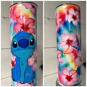 Puede incluir: Un vaso de acero inoxidable con un personaje de dibujos animados azul y blanco con ojos grandes y una gran sonrisa. El vaso tiene un fondo de tie-dye colorido con flores de hibisco rosas.