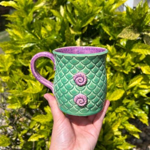 Puede incluir: Una taza de cerámica verde con un diseño de escamas de sirena y dos detalles de conchas marinas rosas. El asa y el borde interior son rosas. La taza se sostiene en una mano sobre un fondo de follaje verde.