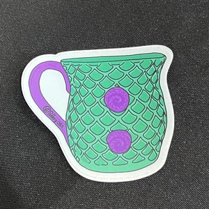 Puede incluir: Una taza de cerámica verde con un asa morada y dos botones morados. La taza tiene un patrón de escamas.
