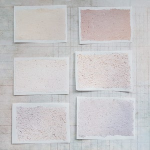 Op de afbeelding: Zes rechthoekige handgemaakte papiersamples in verschillende pastelkleuren, waaronder perzik, roze en lavendel. Elk stuk heeft een getextureerd oppervlak en een gescheurde rand, gerangschikt op een achtergrond met rasterpatroon.