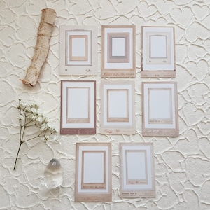 Könnte beinhalten: Acht Fotorahmen im Vintage-Stil mit weißen Zentren und verzierten Rändern in Beige- und Brauntönen. Sie sind auf einer strukturierten weißen Oberfläche angeordnet, mit einem Stück Birkenrinde und Zweigen weißer Blumen.