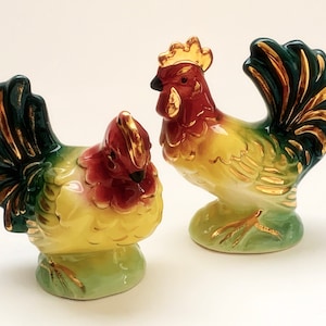 Peut inclure: Deux figurines de coqs en céramique. Les coqs sont peints avec des détails rouges, jaunes, verts et dorés. Ils se tiennent sur une base verte. Les coqs ont des plumes détaillées et une crête dorée.