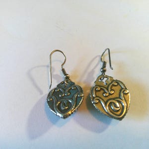 May include: A pair of silver-toned metal drop earrings featuring a heart-shaped pendant with an embossed, swirling filigree pattern. Each earring is attached to a simple curved ear wire hook, showcasing a metallic finish with darkened recessed details.