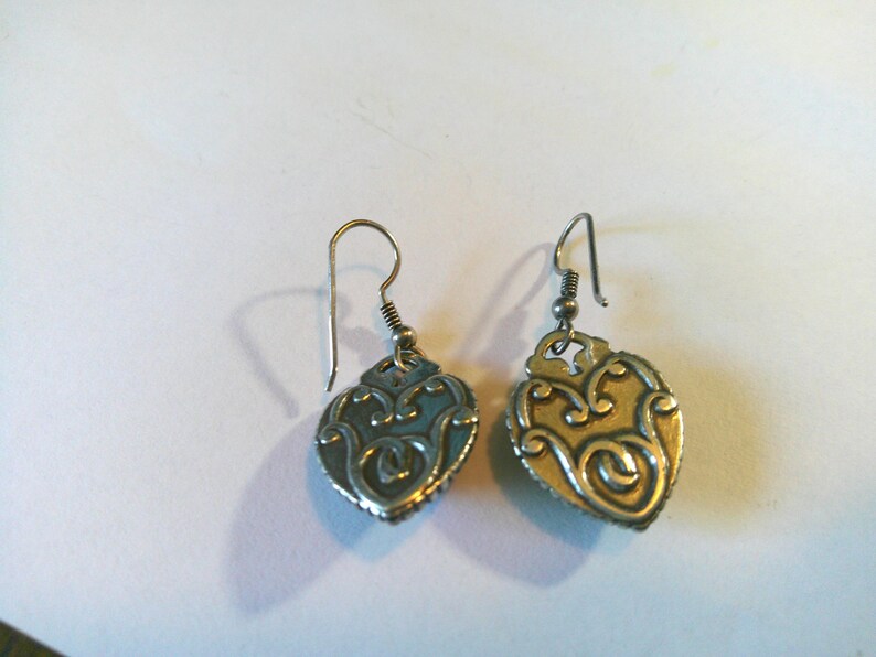 May include: A pair of silver-toned metal drop earrings featuring a heart-shaped pendant with an embossed, swirling filigree pattern. Each earring is attached to a simple curved ear wire hook, showcasing a metallic finish with darkened recessed details.