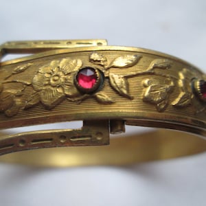 May include: Gold-toned cuff bracelet with floral and leaf engravings. The bracelet features two red gemstones and a clasp closure. The design includes detailed floral patterns and a textured surface, creating a vintage aesthetic.