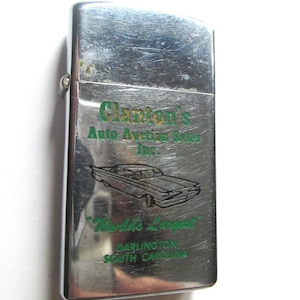 May include: A silver lighter with a green inscription that reads "Clanton's Auto Auction Sales Inc. World's Largest Darlington South Carolina". The lighter has a small car illustration.