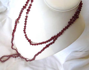 May include: A long, layered necklace with faceted, dark red beads. The necklace is draped over a white bust form, showcasing its length and design. The beads have a sparkling appearance.