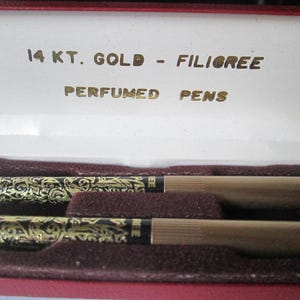 May include: A set of two gold-toned pens with black ink and filigree designs. The pens are in a red velvet box with the words "14 KT. GOLD - FILIGREE PERFUMED PENS" printed in gold.